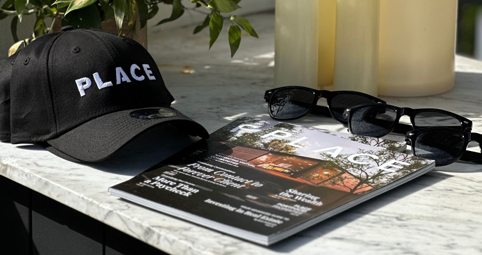 Branded PLACE merchandise including a black hat, magazine, and sunglasses on a marble surface, representing real estate professional lifestyle.