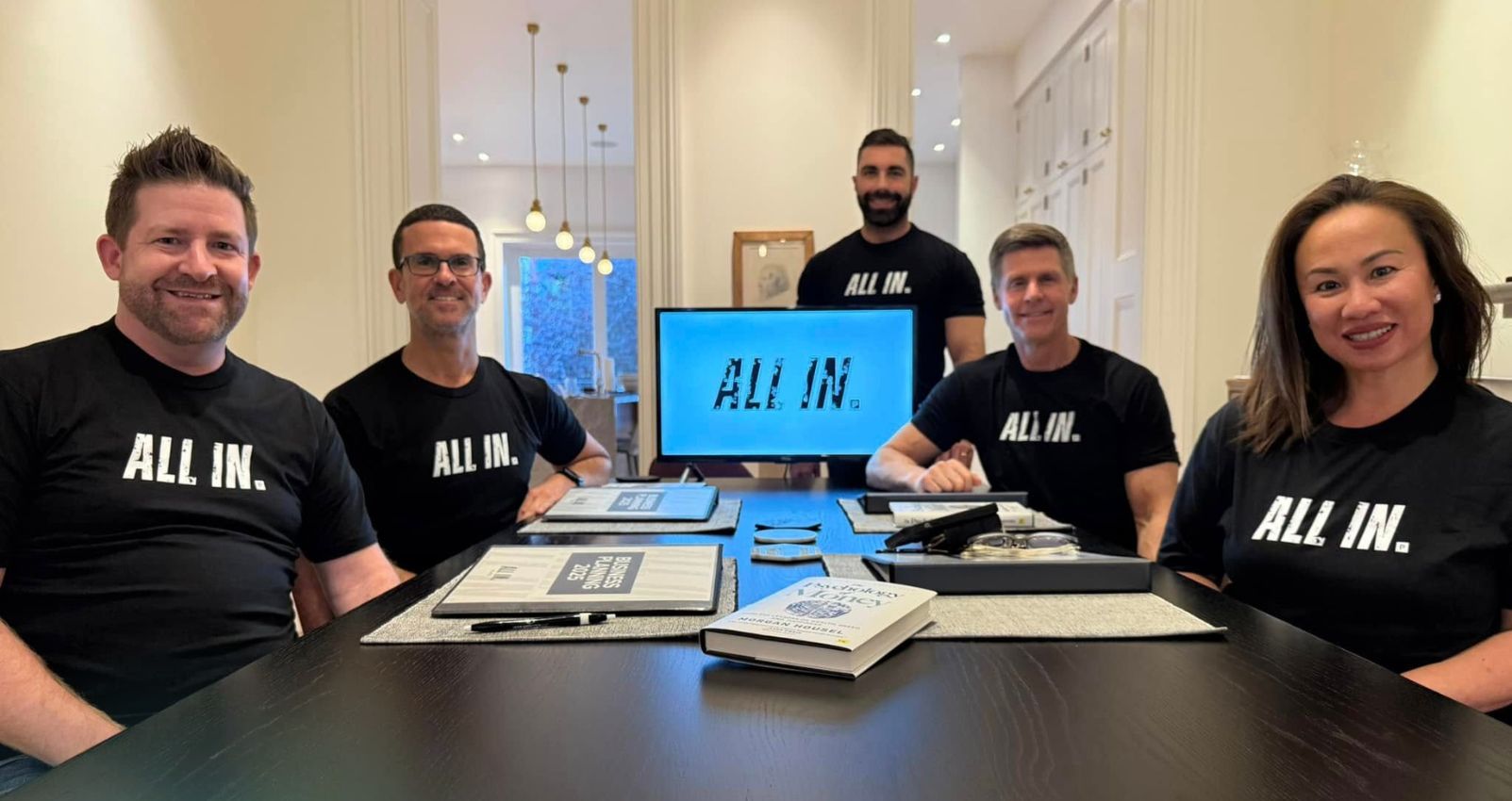 Real estate professionals sitting at a conference table wearing matching "ALL IN." shirts, demonstrating a unified real estate mindset.