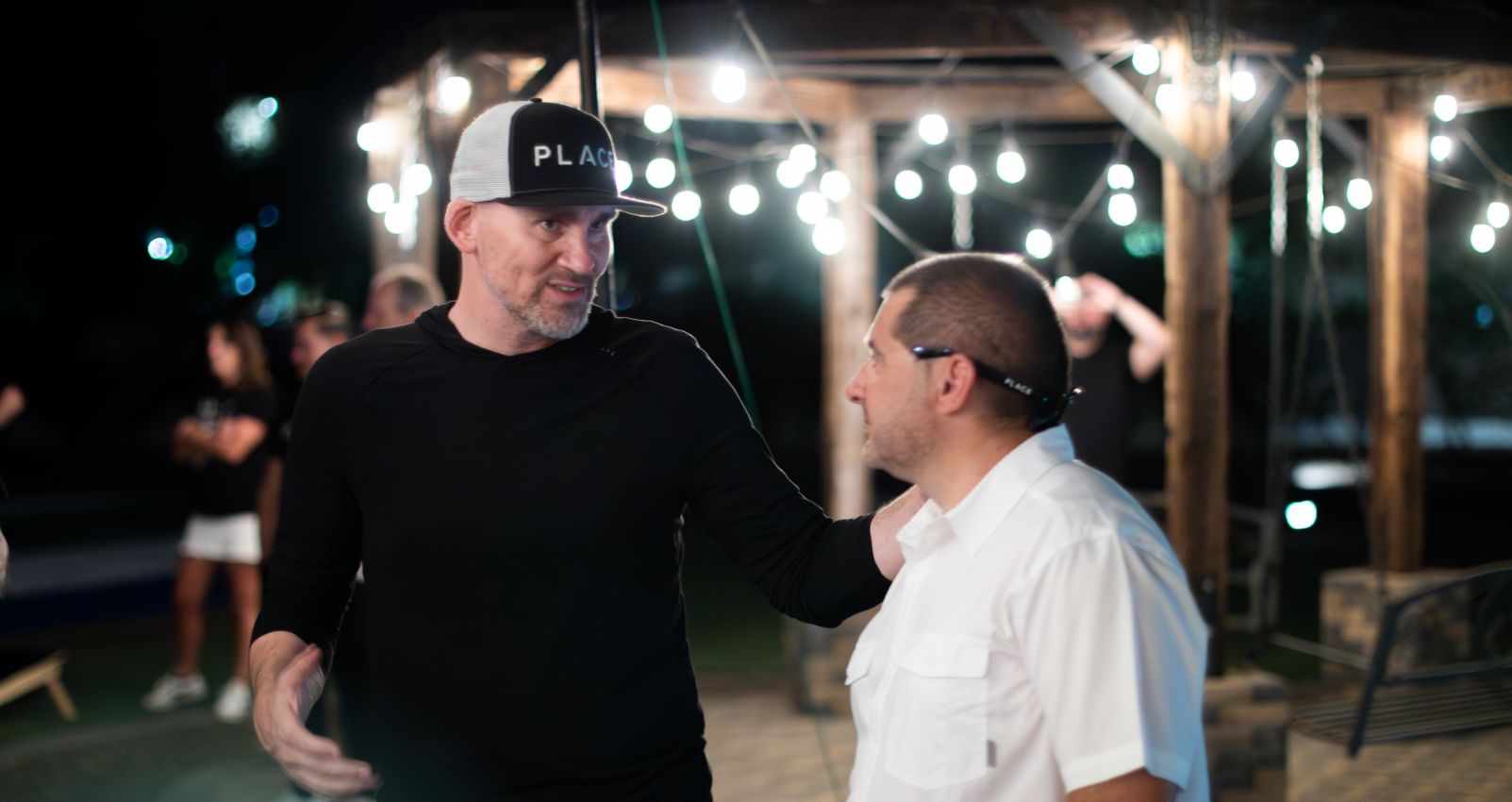 Mastering Influence The 10 Skills Behind Successful Real Estate Sales Two men having a conversation at an outdoor evening event, one wearing a PLACE hat and the other in a white shirt, with string lights hanging in the background.